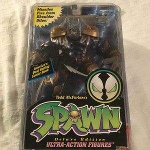 May include: A Spawn action figure in a plastic package. The figure is black and green with gold accents. The figure is holding a sword and has a skull on its chest. The package says "Todd McFarlane's Spawn Deluxe Edition Ultra-Action Figures".