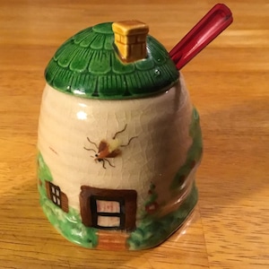 May include: A ceramic honey pot shaped like a cottage, with a green roof and a small chimney. The pot has a beige body with a painted insect and a red spoon. The cottage has a brown door and window, and is surrounded by green foliage.