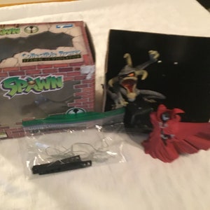 May include: A Spawn collectible figure set with a green, red, and black color scheme. The set includes a Spawn figure, a Spawn figure in a red cape, and a box with a brick wall design. The box has the word "Spawn" printed on it.