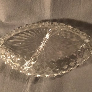 May include: A clear glass divided serving dish with a diamond pattern and a fan-shaped design in the center. The dish has two handles on the sides.
