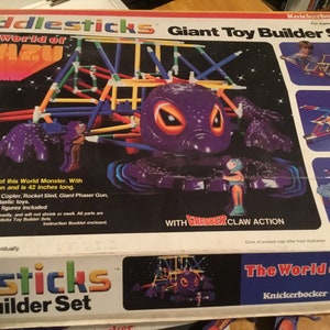May include: Fiddlesticks Giant Toy Builder Set box featuring "The World of Mazu." The box shows a purple monster with grabber claw action, measuring 42 inches long. Includes articulated action figures and various toy builds.