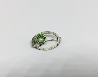 May include: A silver oval brooch with a green clover design and a white pearl in the center.