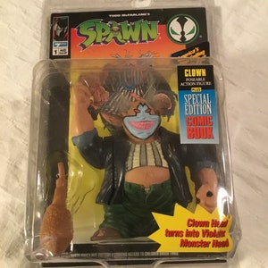 Puede incluir: Una figura de acción de Spawn de un payaso con una cabeza extraíble que se transforma en un monstruo. La figura sostiene un garrote y viene con un cómic. El empaque es verde y amarillo con el logotipo de Spawn.