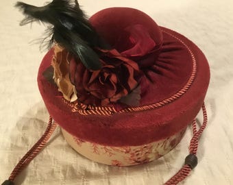 May include: A small, round, red velvet hat with a floral embellishment and a black feather. The hat has a drawstring closure and a cream-colored lining.