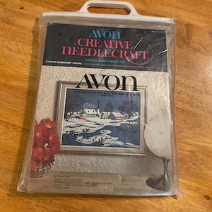 May include: A vintage Avon Creative Needlecraft crewel embroidery kit featuring a winter scene with houses and a river. The kit includes everything needed to complete the project, except for framing. The packaging is white with blue and red accents and features the Avon logo.