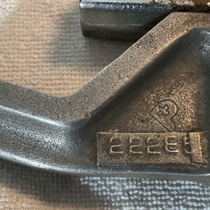 May include: Close-up of a silver-colored metal tool with a curved, hook-like end. The tool has a textured surface and a series of stamped numbers, "22285". A symbol is also stamped on the tool. The tool appears to be a component of a larger machine.