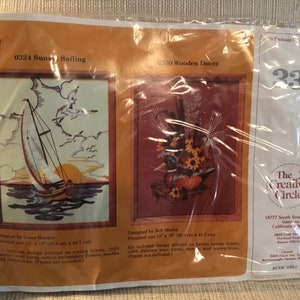 May include: A packaged cross-stitch kit featuring two designs: "Sunset Sailing" with a sailboat and "Wooden Decoy" with a duck and sunflowers. The kit includes printed fabric, yarn, needles, and instructions. Finished sizes are 12" x 16" (30.4 cm x 40.7 cm).
