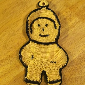 May include: A yellow crocheted pot holder shaped like a person with a smiling face. The pot holder has black trim around the edges.