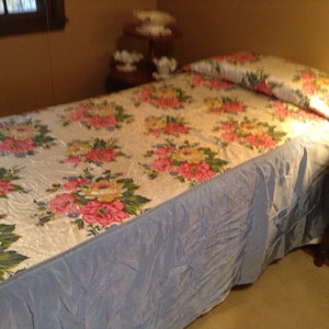 May include: A twin-sized bed with a floral patterned bedspread and a light blue bed skirt. The bedspread features a white background with pink, yellow, and green flowers.