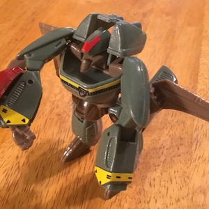 May include: A gray and brown robot toy with red and yellow accents. The robot has wings and is in a standing position.