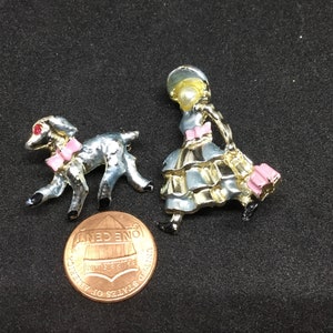 May include: Two vintage enamel pins, one of a girl in a blue dress and hat holding a pink bag, and the other of a gray lamb with a pink bow.