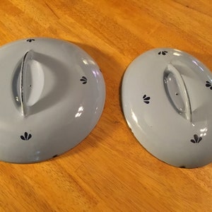 May include: Two light blue enamel lids with black floral designs and a handle. The lids are on a wooden surface.