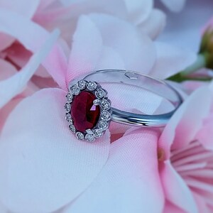 May include: A silver ring with a large oval ruby center stone, surrounded by a halo of small diamonds. The ring is set against a backdrop of pink and white flower petals, creating a soft, romantic aesthetic.