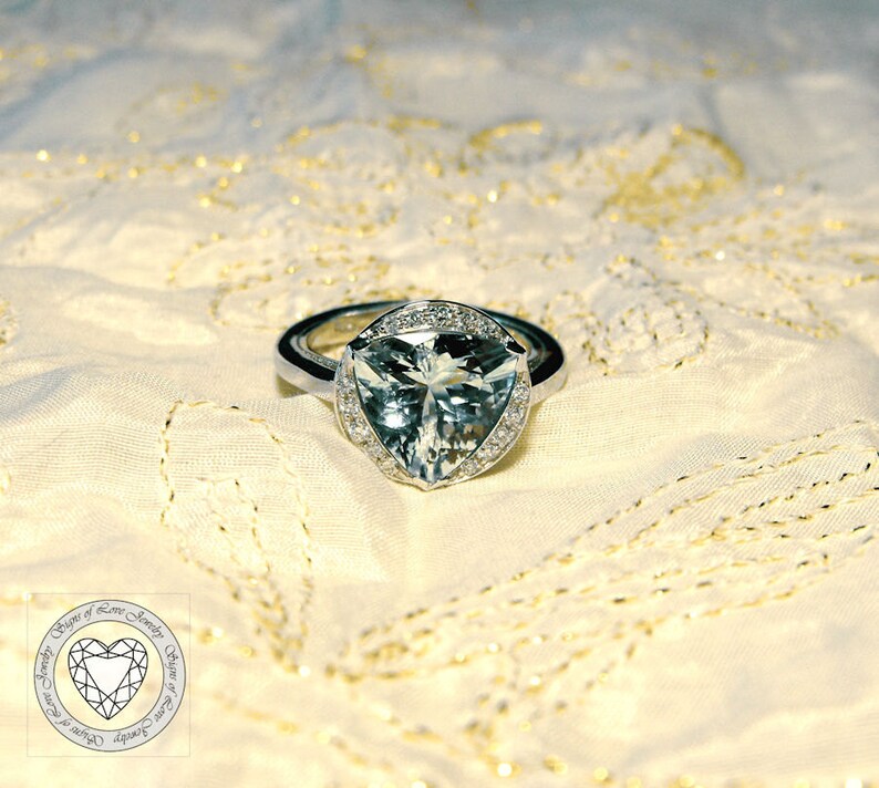 May include: A silver ring with a large, triangular, clear aquamarine gemstone surrounded by smaller diamonds. The ring is on a white, textured fabric background.