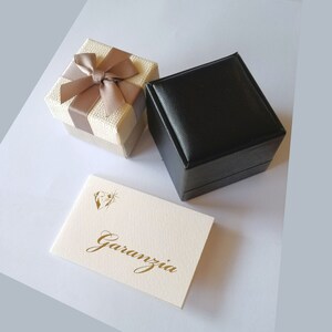 May include: Two gift boxes, one beige with a brown bow and one black, with a white card that says "Garanzia" in gold lettering.