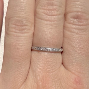 May include: A delicate silver ring with a band of small, sparkling stones. The ring is set on a hand, showcasing its fine details and elegant design. The ring's simple yet sophisticated style makes it a versatile piece of jewelry.