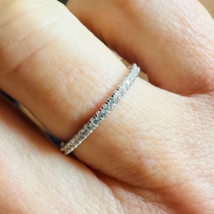 May include: A delicate, thin ring with a band of small, clear gemstones. The ring is set in a silver or white metal and is worn on a finger. The gemstones sparkle, catching the light.