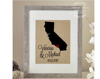 Puede incluir: Una impresión enmarcada en tela de arpillera con una silueta negra del estado de California con un corazón rojo marcando una ubicación. La impresión incluye los nombres "Vanessa & Michael" y la fecha "10.15.2017".