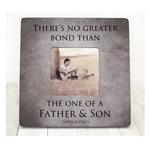 Puede incluir: Un marco de fotos gris con una inscripción negra que dice "There's no greater bond than the one of a father & son. Daddy & Ethan". El marco tiene una foto de un padre y un hijo sentados en un campo, el padre está tocando la guitarra.