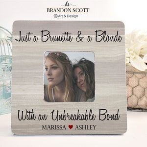 May include: A light wood picture frame with a photo of two women, one with brown hair and one with blonde hair. The frame has the text "Just a Brunette & a Blonde" at the top and "With an Unbreakable Bond" at the bottom. The names "MARISSA" and "ASHLEY" are written below the text, with a red heart symbol between them.