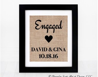 Puede incluir: Una impresión de arpillera enmarcada con el texto "Engaged" en fuente cursiva negra, un corazón negro y los nombres "David & Gina" y la fecha "10.18.16" en texto negro.