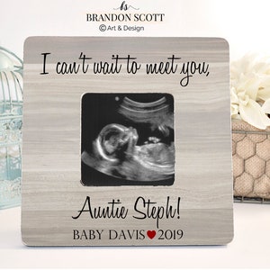 May include: A grey wooden picture frame with a white mount and a black and white ultrasound image inside. The frame has the text "I can't wait to meet you, Auntie Steph! Baby Davis 2019" printed on it.