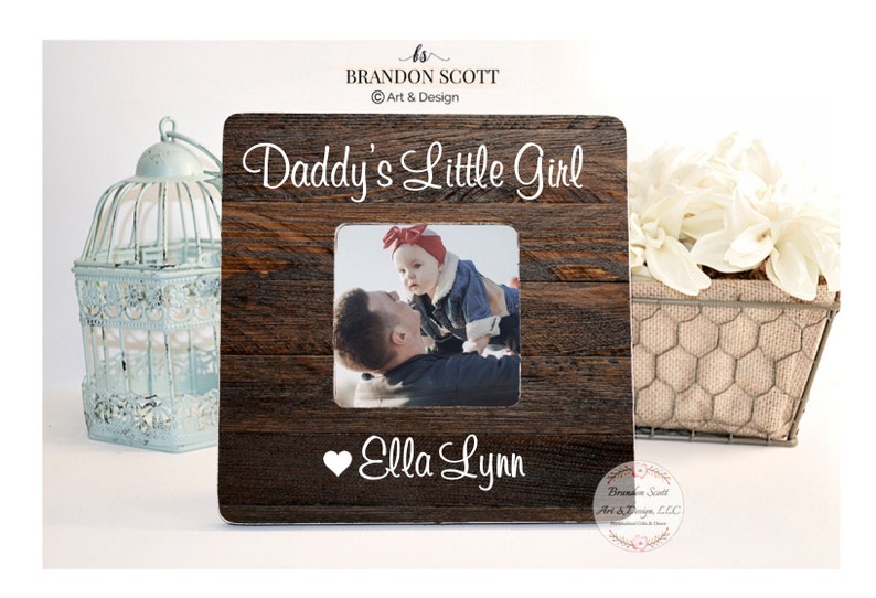 May include: A rustic wooden picture frame with a white inscription that reads "Daddy's Little Girl" and a heart with the name "Ella Lynn". A photo of a father holding his daughter is in the frame.