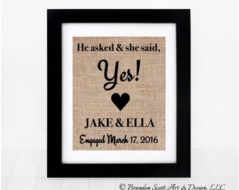 Puede incluir: Una impresión de arpillera enmarcada con texto negro que dice "He asked & she said, Yes!" con un corazón y los nombres "JAKE & ELLA" y la fecha "Engaged March 17, 2016".
