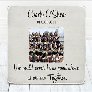 May include: A white wooden picture frame with a black inscription that reads "Coach O'Shea #1 COACH". The frame holds a photo of a cheerleading squad in their uniforms. The inscription below the photo reads "We could never be as good alone as we are Together."