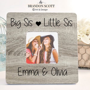 May include: Square wooden picture frame with a gray wood grain finish. The frame has the words "Big Sis ♥ Little Sis" and the names "Emma & Olivia" printed on it. The frame holds a photo of two smiling women.