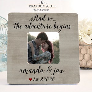 May include: A light brown wooden picture frame with a white mat and a photo of a couple. The frame has the text "And so... the adventure begins" at the top and "amanda & jax Est. 2.20.20" at the bottom.