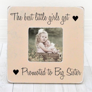 May include: A light pink wooden picture frame with the text "The best little girls get ♥ Promoted to Big Sister" and a black heart on each side of the text. The frame has a photo of two young girls in it.