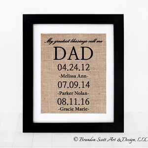May include: A framed burlap print with the word "DAD" and dates of birth for three children. The text reads "My greatest blessings call me DAD 04.24.12 -Melissa Ann- 07.09.14 -Parker Nolan- 08.11.16 -Gracie Marie-  Brandon Scott Art & Design, LLC"