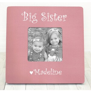May include: Pink wooden picture frame with the text "Big Sister" and a heart with the name "Madeline" below the picture. The frame has a black and white photo of two young girls.