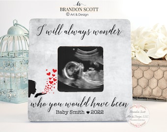 May include: A white picture frame with a gray background and black text that reads "I will always wonder who you would have been Baby Smith 2022". The frame has a black silhouette of a woman with red hearts coming out of her hand. There is a black and white ultrasound image in the frame.