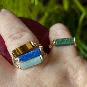 Puede incluir: Tres anillos de oro con piedras de diferentes colores. El anillo superior tiene una piedra de ojo de tigre, el anillo del medio tiene una piedra de lapisl&aacute;zuli azul y el anillo inferior tiene una piedra de aventurina verde.