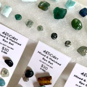 May include: Assortment of polished gemstones in various colors, including green, blue, and brown. Some stones are displayed on small cards with the text "ARTICINDY Mixed & Matched 8pc Set". The gemstones vary in size and shape.