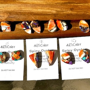 May include: Four pairs of Spiny Oyster earrings, each pair featuring a unique combination of orange, red, white, purple, and turquoise colors. The earrings are displayed on white cards with the text "ARTICINDY Spiny Oyster hope courage creativity Do NOT Get Wet".