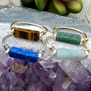 May include: Four gold rings with different colored stones. The stones are a tiger's eye, a green stone, a blue stone, and a light green stone.