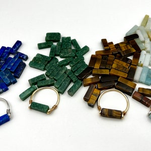 Puede incluir: Cuatro anillos con piedras preciosas de diferentes colores. Los anillos est&aacute;n hechos de alambre de plata y oro y presentan una sola piedra preciosa en el centro. Las piedras preciosas son lapisl&aacute;zuli, aventurina verde, ojo de tigre y aguamarina.