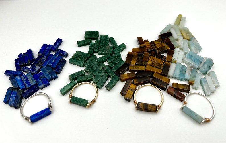 Puede incluir: Cuatro anillos con piedras preciosas de diferentes colores. Los anillos est&aacute;n hechos de alambre de plata y oro y presentan una sola piedra preciosa en el centro. Las piedras preciosas son lapisl&aacute;zuli, aventurina verde, ojo de tigre y aguamarina.