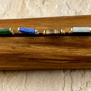 Puede incluir: Cuatro anillos de piedras preciosas diferentes en un expositor de madera. Los anillos presentan piedras preciosas verdes, azules, marrones y azul claro.