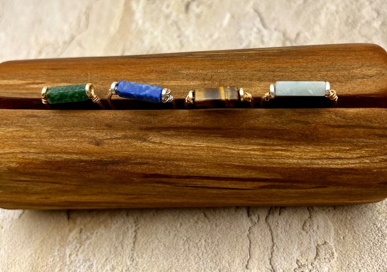 Puede incluir: Cuatro anillos de piedras preciosas diferentes en un expositor de madera. Los anillos presentan piedras preciosas verdes, azules, marrones y azul claro.