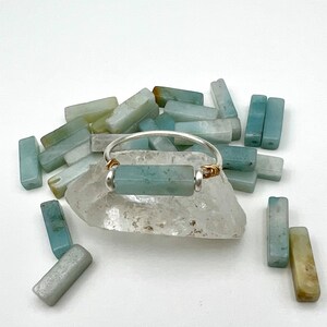 Puede incluir: Un anillo de plata con una piedra rectangular de color azul claro engastada en el centro. El anillo est&aacute; rodeado de una dispersi&oacute;n de piedras rectangulares de color azul claro y verde.