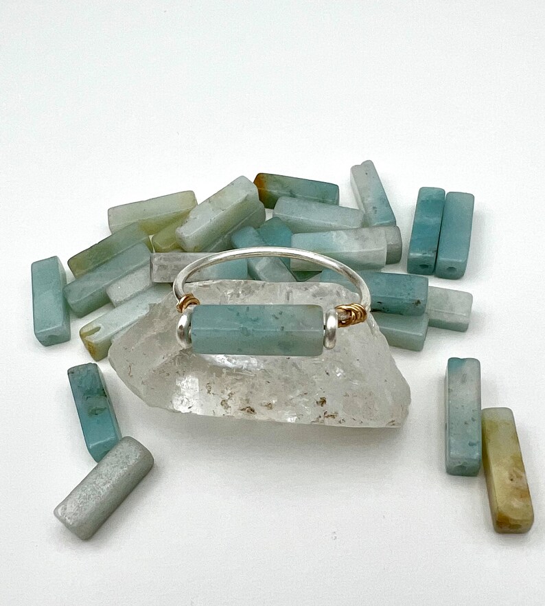 Puede incluir: Un anillo de plata con una piedra rectangular de color azul claro engastada en el centro. El anillo est&aacute; rodeado de una dispersi&oacute;n de piedras rectangulares de color azul claro y verde.