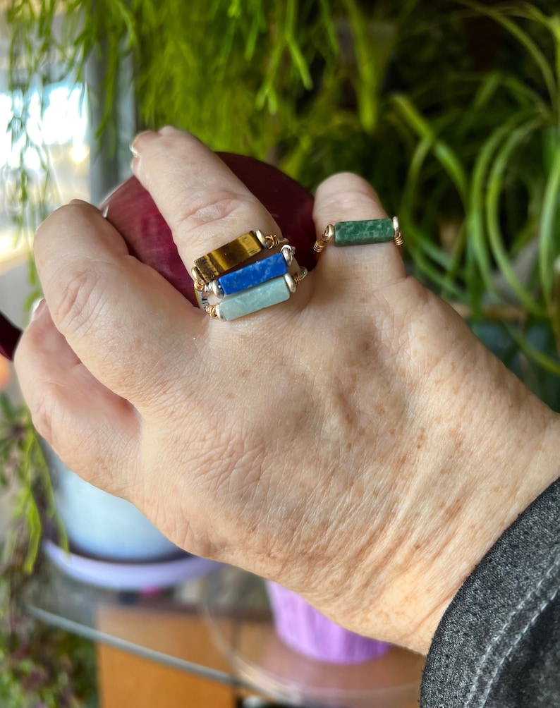 Puede incluir: Tres anillos de oro con piedras de diferentes colores. El anillo superior tiene una piedra de ojo de tigre, el anillo del medio tiene una piedra de lapisl&aacute;zuli azul y el anillo inferior tiene una piedra de aventurina verde.