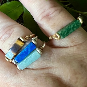 Puede incluir: Cuatro anillos de alambre dorado con piedras de diferentes colores. Los anillos est&aacute;n apilados en un dedo. Las piedras son verdes, azules, azul claro y marrones.