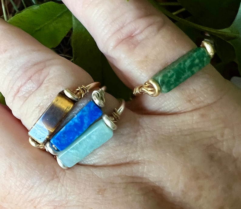Puede incluir: Cuatro anillos de alambre dorado con piedras de diferentes colores. Los anillos est&aacute;n apilados en un dedo. Las piedras son verdes, azules, azul claro y marrones.