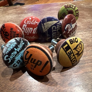 May include: Eight vintage-style cabinet knobs with various colorful designs and text, including "Gold-Sun-Drop", "Snow White", "Yup!", "Bulls Eye", "Frostie", and "Big Lemon".