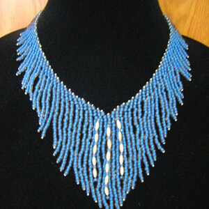 May include: A blue and white beaded necklace with a fringe design. The necklace is displayed on a black mannequin bust.
